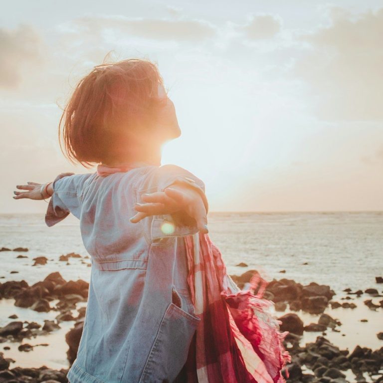 „Wenn der See still wird, erkennst du dich selbst – Coaching in der Natur.“ Eine Frau mit ausgebreiteten Armen steht am Strand bei Sonnenuntergang und erlebt die Kraft der Sonne.